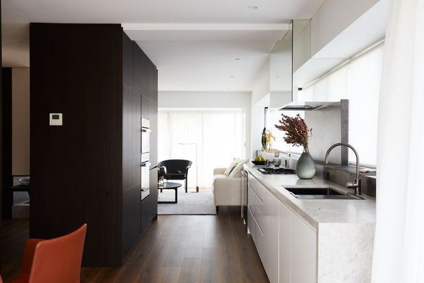 A bright contemporary open-plan kitchen viewed toward an adjoining living area. The kitchen features dark timber-toned full-height cabinetry, white lower cabinetry, a pale stone benchtop with integrated sink, stainless steel appliances and a curved metal tap. Floor-to-ceiling glazing and sheer curtains bring in abundant natural light, while a dining or living zone is visible beyond.