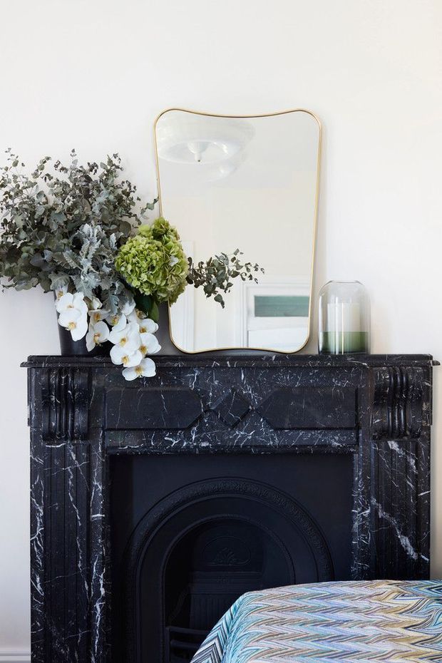 A close-up interior vignette centred on an ornate black marble fireplace with carved detailing and a dark firebox. A large gold-framed mirror, floral arrangement, and glass decorative vessel sit on the mantel, while the corner of a patterned upholstered piece appears in the foreground. The room has white walls and a refined, heritage-inspired character.