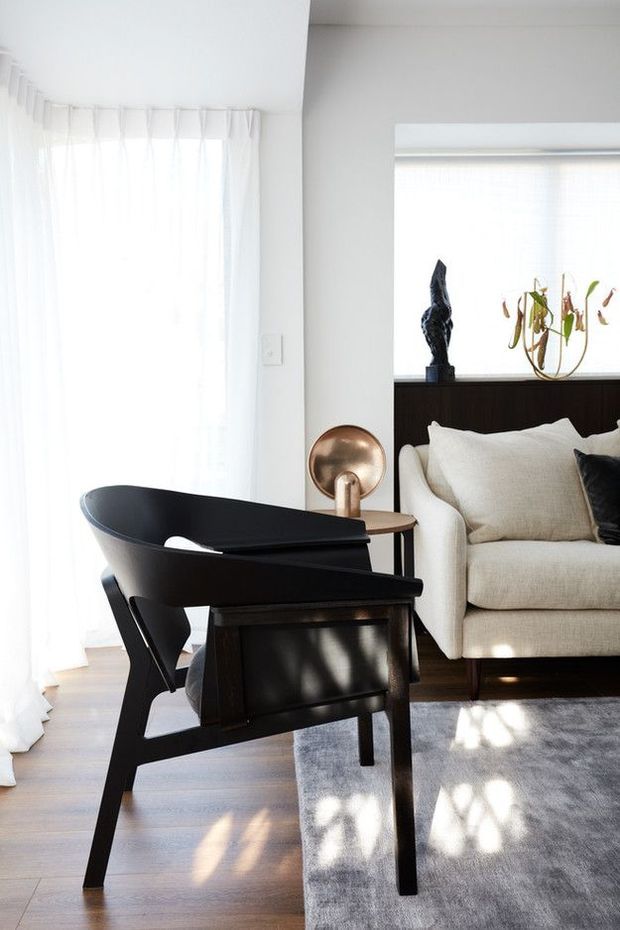 Bright, refined living room with sheer full-height curtains, pale walls, light timber flooring and a soft grey rug. A sculptural black leather lounge chair and dark wood side table sit in the foreground, beside a cream upholstered sofa with neutral cushions. A bronze-toned circular lamp, dark timber cabinetry, artwork and indoor plants add restrained accents. Strong natural light creates a calm, contemporary atmosphere.