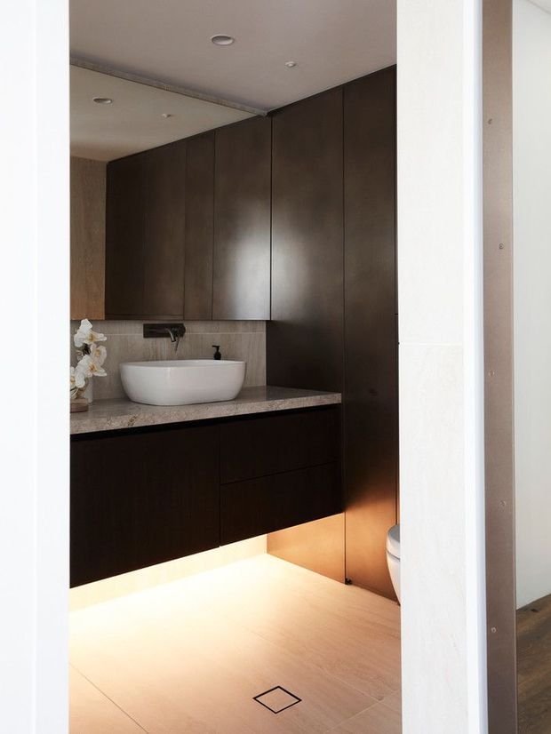A compact contemporary bathroom viewed through an open doorway, featuring dark floor-to-ceiling timber cabinetry, a pale stone benchtop, a white vessel basin, wall-mounted tapware, tiled walls and floor, and warm under-cabinet lighting. A partial toilet is visible at the right.