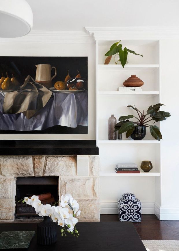 A refined living room vignette centered on a traditional stone fireplace with a black mantel and framed still-life artwork. Built-in white shelving displays potted plants, ceramics, books, and decorative objects. A dark coffee table, floral arrangement, and hardwood flooring complete the warm, curated interior.