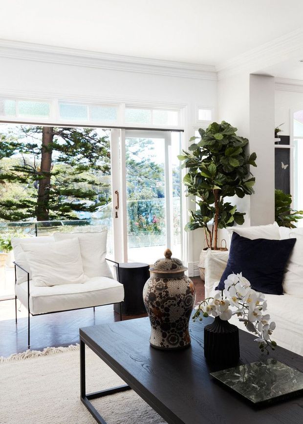 Bright, refined living room with white walls, ornate ceiling cornices and trim, large timber-framed windows and glazed doors, and dark polished timber flooring. The room is furnished with a white upholstered armchair and sofa, navy accent cushions, a black rectangular coffee table, decorative ceramic vessel, and potted fiddle-leaf fig. Lush greenery is visible outside through the glazing.