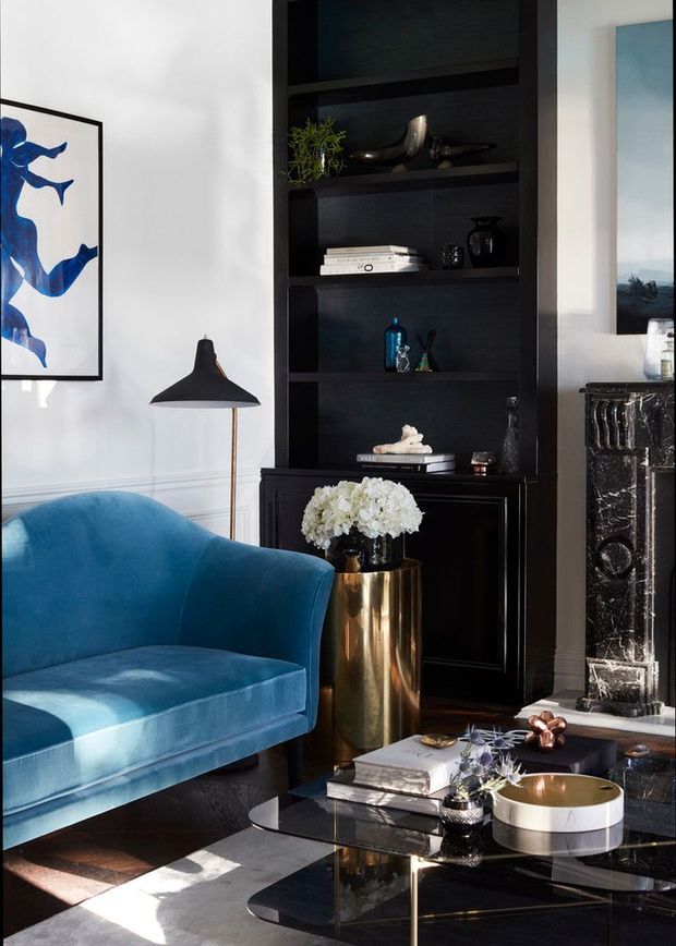 Refined living room vignette featuring a deep blue velvet armchair, black built-in shelving and cabinetry, a black marble fireplace surround, brass planter, black floor lamp, artwork, and a glossy black coffee table. The scheme combines strong blue accents with black, white, and metallic details in a polished, high-contrast setting.
