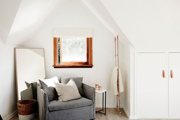 A bright, compact attic-style living nook with a grey upholstered armchair, neutral cushions, a small side table, woven basket, leaning mirror, timber-framed window, and built-in white storage cupboards. The white pitched ceiling and warm timber details create a calm, airy interior.
