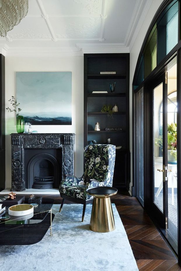 Elegant traditional living room with a black marble fireplace, ornate white coffered ceiling and mouldings, dark built-in shelving, arched black-framed glazed doors, a floral upholstered armchair, brass side table, and dark herringbone timber flooring. A pale blue rug and abstract artwork add softer colour to the refined space.