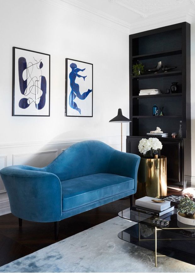 Refined living room featuring a deep blue velvet two-seat sofa, black built-in shelving, white decorative wall panelling and ornate ceiling cornices. Abstract blue artwork, a black floor lamp, a brass cylindrical side table, and a layered glass coffee table complete the sophisticated interior.