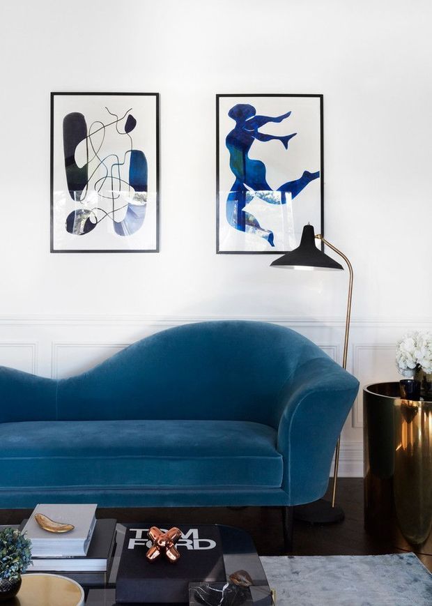 A refined living room with a deep blue curved velvet sofa, white walls with decorative panel moulding, two blue-toned abstract artworks in black frames, and a sculptural black-and-brass floor lamp. A glossy dark side table, coffee table with books and decor, and a pale rug complete the polished interior.