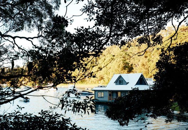 A small waterfront house viewed through dense tree branches, with a steep pitched gabled roof, dark horizontal cladding, and multiple windows. The house sits directly beside calm water, with a wooded hillside behind it. The image has a scenic, secluded coastal or lakeside character.