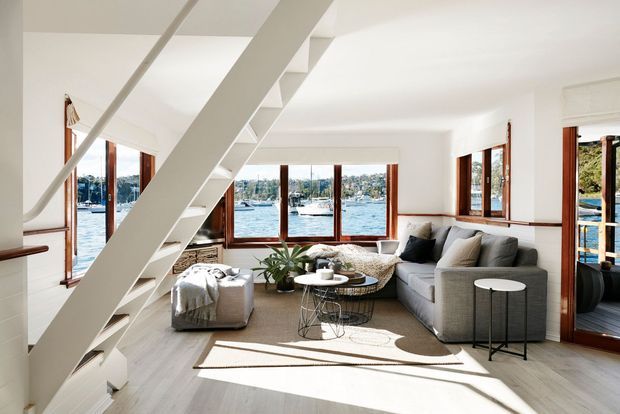 Bright, contemporary waterfront living room with large timber-framed windows and glazed doors overlooking a marina. The room features a grey sectional sofa, round coffee tables, an upholstered ottoman, a potted plant, pale timber flooring, white walls, and a striking open white staircase with integrated-looking treads.