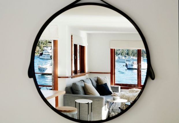 A round black-framed wall mirror reflects a bright waterfront living room with white walls, timber-trimmed windows, a grey sofa, small round tables, and boats visible outside on the water. The image is primarily focused on the mirror and the reflected interior space.