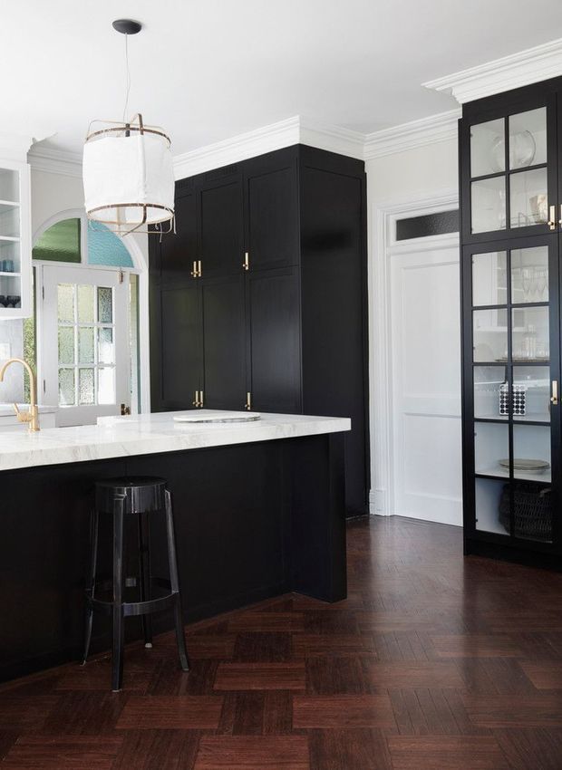 A refined, traditional kitchen featuring a large black island with a white stone benchtop, tall black cabinetry, glass-front display storage, white panelled doors and walls, decorative crown moulding, and dark herringbone timber flooring. Brass hardware and a suspended fabric pendant add warm accents.