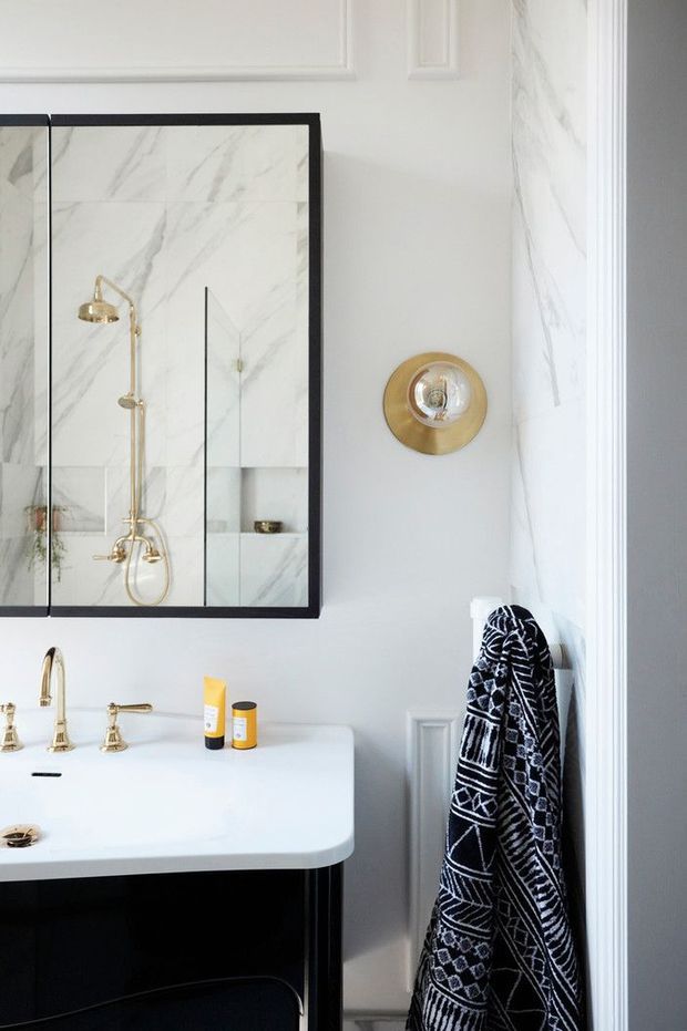 A refined bathroom vanity area with a white stone countertop, black cabinetry, brass tapware, and a black-framed glass shower enclosure. Marble-look wall surfaces are visible inside the shower, while pale grey walls feature decorative raised moulding. A patterned dark towel hangs beside the vanity, and a brass wall light adds a warm accent.