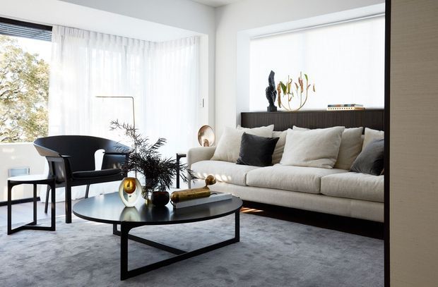 Bright, refined living room with floor-to-ceiling corner glazing dressed in sheer white curtains, a large cream sectional sofa, black accent cushions, a dark round coffee table, sculptural decor, and a grey area rug. The room has a clean contemporary shell with strong mid-century-inspired furniture and restrained monochrome styling.