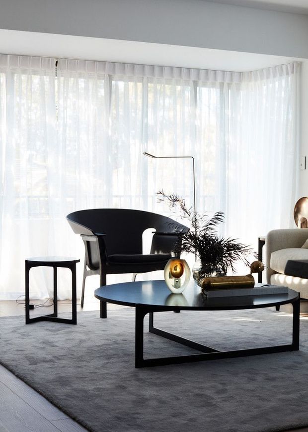 Bright, refined living room with floor-to-ceiling sheer curtains, a black sculptural lounge chair, low black rectangular coffee table, pale upholstered sofa, and minimalist decorative objects arranged over a large grey rug. The space has abundant filtered daylight and a clean, contemporary feel.