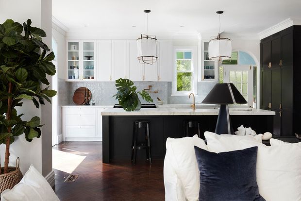 Bright open-plan kitchen viewed from the living area, featuring a large black island with a pale stone benchtop, white built-in cabinetry and shelving, dark timber flooring, pendant lights, and a black-framed window and door zone. A white sofa with dark blue cushions occupies the foreground, while leafy indoor plants add greenery. The room has a refined, high-contrast traditional-contemporary character with abundant natural light.