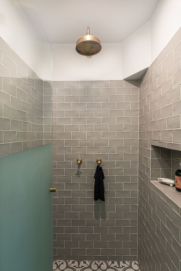 Compact walk-in shower with grey subway wall tiles, white painted ceiling, patterned encaustic-style floor tiles, a partial frosted green-glass door, brass shower fitting and hooks, and a recessed tiled shelf with toiletries. The space has a simple contemporary design with warm metallic detailing.