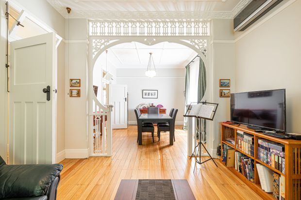 Bright traditional interior view through an ornate decorative archway, with a painted timber door, polished timber floorboards, dining table and chairs, television and timber media cabinet, and a glimpse of a staircase beyond. The room features detailed ceiling cornices, lattice-style arch detailing, and warm natural light.