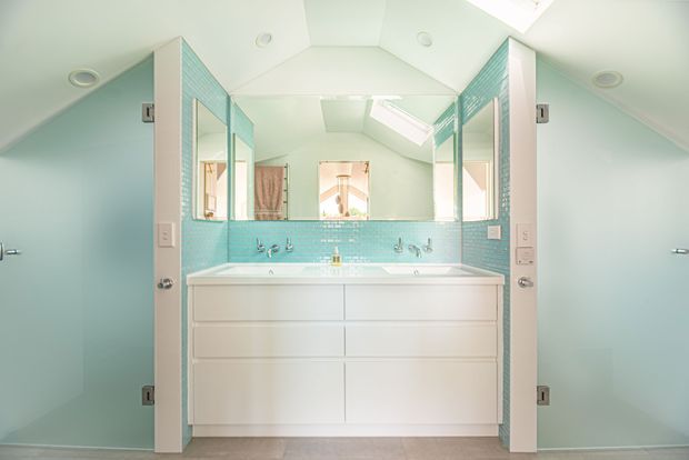 Bright, symmetrical bathroom with a large white double vanity and integrated basins, pale aqua wall paint, turquoise small-format tile splashback, mirrored cabinet doors, skylights, and a vaulted ceiling. The room has a clean, airy contemporary coastal feel.