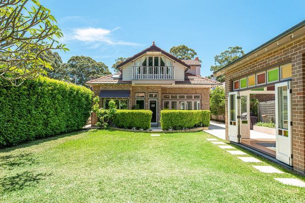 Sunny exterior view of a traditional brick house with a terracotta-tiled gabled roof, upper-level balcony, symmetrical multi-pane windows, and manicured hedges. A contemporary brick extension with glazed bi-fold doors and colourful transom panels opens onto the broad lawn and stepping-stone path.