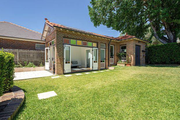 A single-storey brick house extension viewed from a landscaped backyard. The addition has a low-pitched tiled roof, large glazed sliding doors, and a colourful strip of small rectangular glazing above the openings. A paved path, lawn, established trees, hedges, and a dark brick outdoor structure frame the scene.
