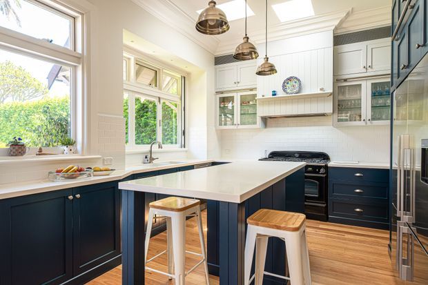 Bright, spacious kitchen with a large central island, dark navy lower cabinetry, white shaker-style upper cabinets, timber flooring, white subway-tile splashback, and pendant lighting. The room features a classic renovated character-home feel with abundant natural light, a freestanding-style range cooker, stools at the island, and garden views through timber-framed windows.