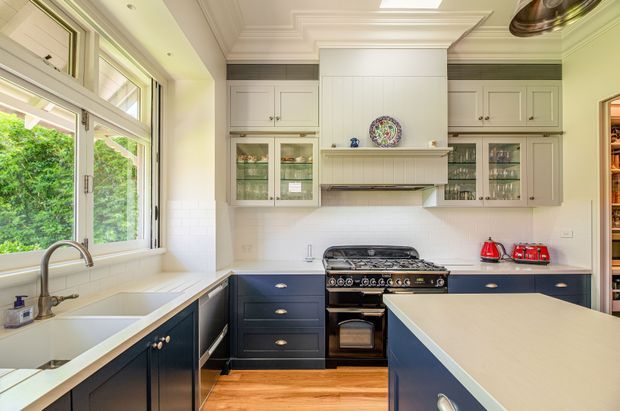 Bright, traditional-style kitchen with dark navy lower cabinetry, white upper cabinets, glass-front display cupboards, a large central island, freestanding range cooker, farmhouse-style sink, and warm timber flooring. The room features detailed cornices, a coffered ceiling treatment, marble-look benchtops, and large windows overlooking greenery.