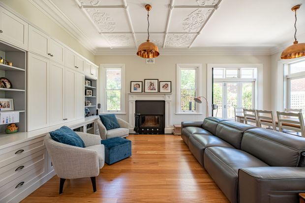 A bright, traditionally detailed open-plan living room with built-in cabinetry along one wall, a central fireplace, upholstered armchairs, a large dark leather sectional, and a dining area by the windows. The room features polished timber flooring, decorative pressed ceiling panels, ornate cornices, white-painted joinery, and warm copper pendant lights.