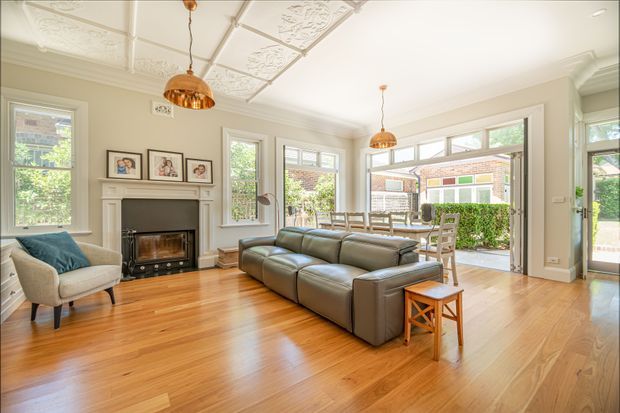 Bright, spacious traditional living room with polished timber flooring, a grey sectional sofa, fireplace with mantel, framed artwork, decorative pressed ceiling panels, pendant lights, and large windows and glazed doors opening to a hedge-lined outdoor area.