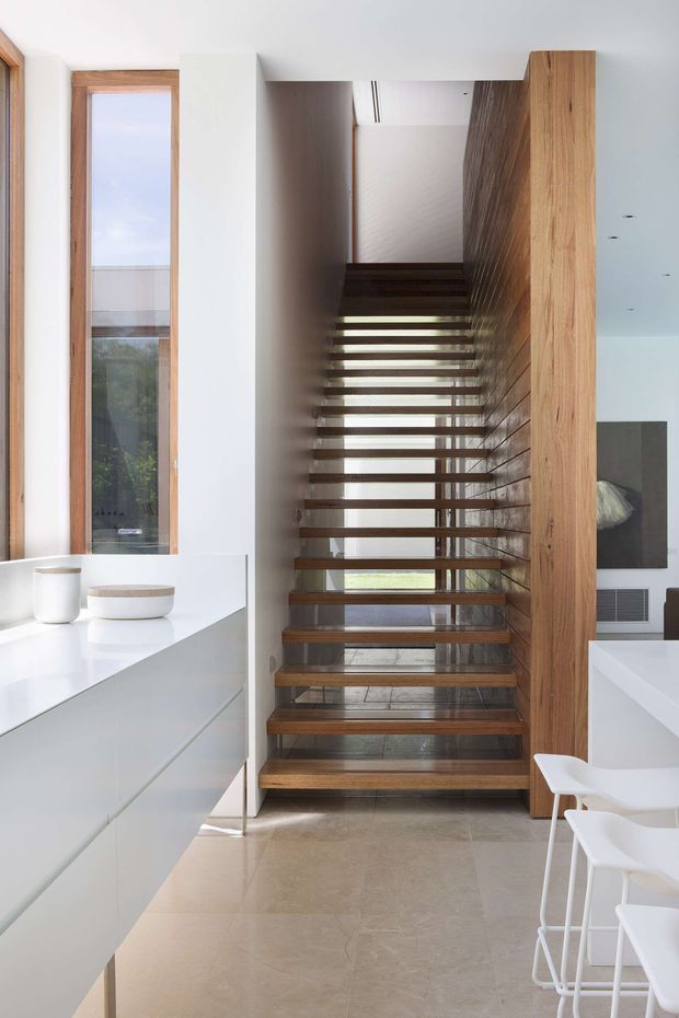 A bright, minimalist contemporary interior focused on an open-riser timber staircase. The staircase has dark timber treads, slim metal supports, and a full-height timber-lined wall, with a narrow timber-framed window beside it. White built-in cabinetry and a white countertop occupy the foreground, accompanied by white stools. Large light stone or concrete-look floor tiles and white walls create a clean, airy composition.