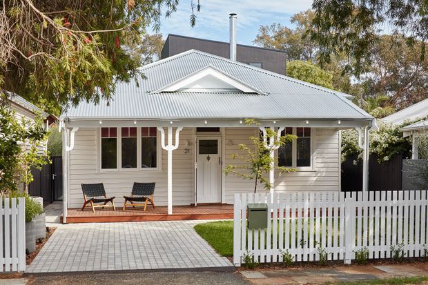 Front exterior view of a compact single-storey weatherboard house with a light grey corrugated pitched roof, decorative gable detail, covered entry porch, white trim, timber decking, paved driveway, and white picket fencing. Mature trees partially frame the suburban streetscape.