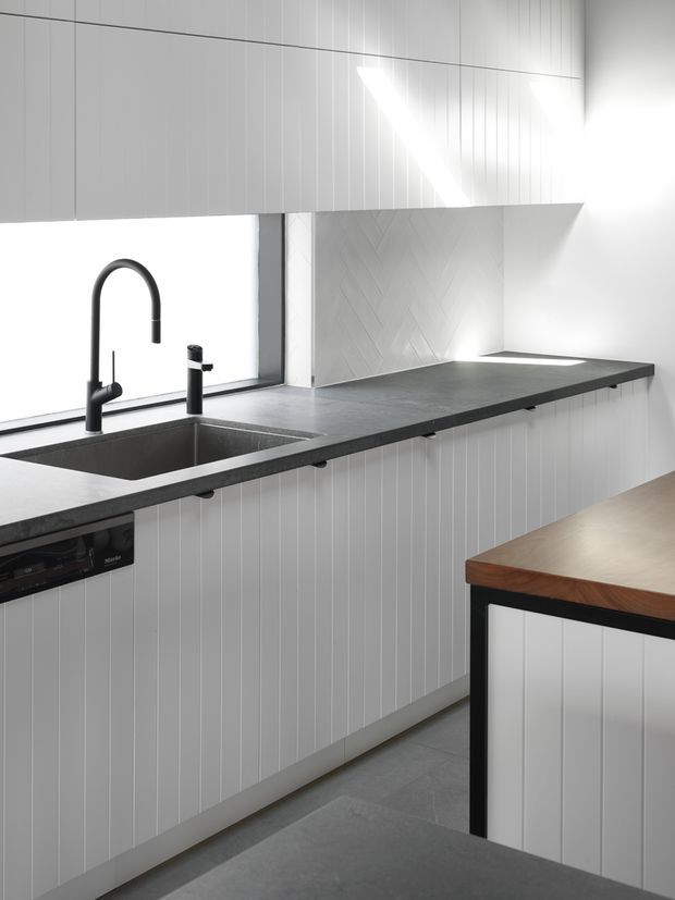 A bright, minimalist contemporary kitchen with white vertically panelled cabinetry, dark charcoal benchtops, an integrated double sink and matte black gooseneck tapware. The backsplash features pale herringbone-patterned tiles, while a timber-topped island or dining bench with black framing occupies the foreground. Strong natural light enters through the large window, creating a clean, high-contrast interior.