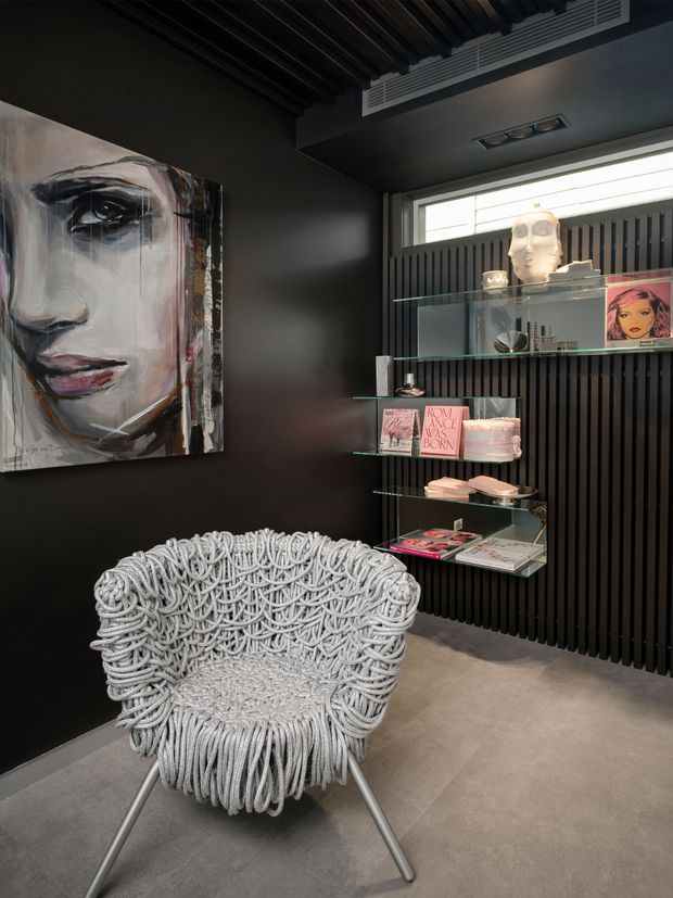 A dark, art-focused interior featuring a large expressive portrait painting, a sculptural white woven lounge chair, and floating glass display shelves holding books, magazines, and decorative objects. The walls and ceiling are charcoal, with a black vertical slatted feature wall, concealed linear ventilation, recessed lighting, and a large horizontal window. The floor is a smooth grey concrete-like finish. The overall atmosphere is dramatic, curated, and gallery-like.