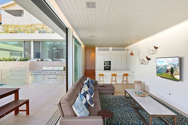 Bright contemporary open-plan living room with a compact kitchen visible at the rear and a large glazed opening onto a paved outdoor entertaining area with a built-in barbecue. The room features a long beige sectional sofa with patterned cushions, patterned area rug, timber coffee table, white built-in kitchen cabinetry, timber accents, bar stools, and a wall-mounted television. The ceiling has white linear panelling and the glazing creates a strong indoor-outdoor connection.
