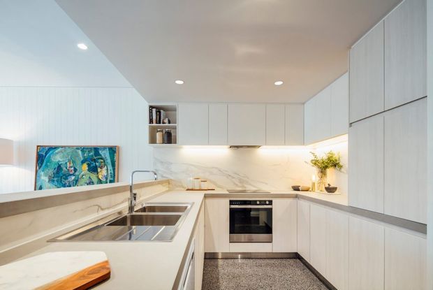 Bright contemporary galley-style kitchen with extensive pale built-in cabinetry, a white marble-look benchtop and splashback, integrated stainless-steel sink and tap, built-in oven and cooktop, under-cabinet lighting, and a terrazzo-like grey floor. A small open shelf and artwork add subtle visual interest.