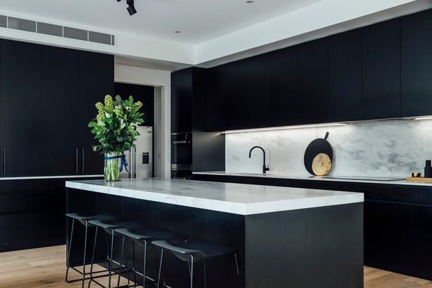 Contemporary kitchen with a large central island, dark matte cabinetry, pale veined stone benchtops and splashback, integrated appliances, and slim black bar stools. Light timber flooring and under-cabinet lighting provide contrast, while a vase and decorative objects add restrained styling.