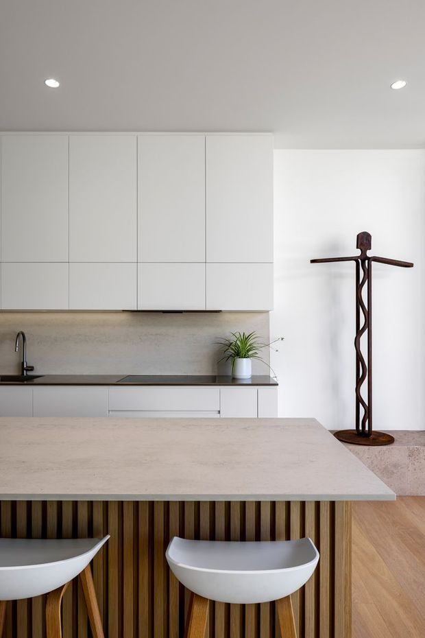A bright, minimalist contemporary kitchen with a large pale stone island and integrated seating. The island front has vertical dark timber slats, contrasting with white curved bar stools. The rear wall features full-height white handleless cabinetry, a dark benchtop, pale textured splashback, integrated sink and cooktop, and a small potted plant. Light timber flooring and a sculptural dark wood figure add warmth and visual interest.
