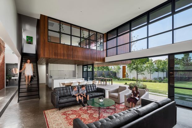 A spacious double-height contemporary living room with full-height glazed walls opening to a landscaped garden. The room features black leather sofas, light upholstered armchairs, a patterned area rug, a glass coffee table, polished concrete flooring, and a dramatic timber-clad mezzanine or upper-level bridge with black-framed glazing. An open stair and dining area are also visible.