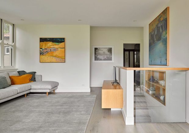 Bright contemporary living room with a light grey sectional sofa, large neutral rug, pale timber flooring, and a white built-in entertainment/storage unit with glazed shelving. Abstract artwork adds warm orange and blue accents, while a partial hallway and stair or landing area are visible beyond the room.