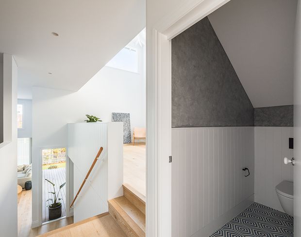 Bright contemporary stair landing viewed beside a compact bathroom. The landing has pale timber stairs and flooring, white walls, a skylight, and a white vertical-panel balustrade with a timber handrail. The bathroom features dark grey textured upper walls, white vertical wall panelling, a patterned geometric floor, and a visible toilet.