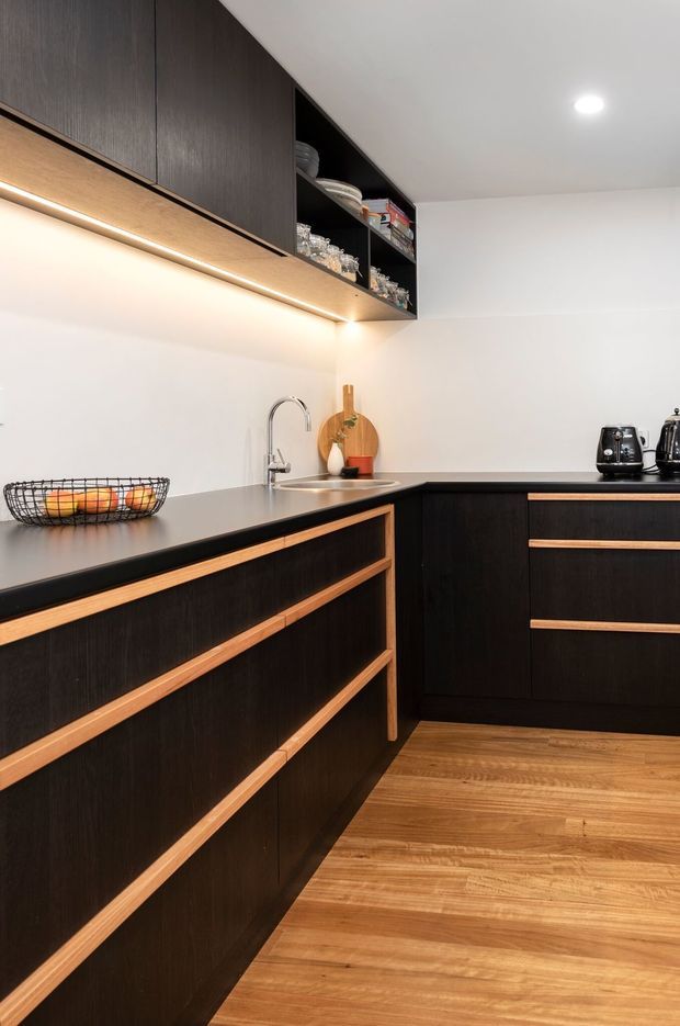 A modern L-shaped kitchen with dark timber cabinetry accented by light timber drawer pulls and trim. The kitchen features a dark benchtop, white walls and splashback, integrated open shelving with under-cabinet lighting, a stainless-steel sink and curved tap, small appliances, and warm timber flooring.