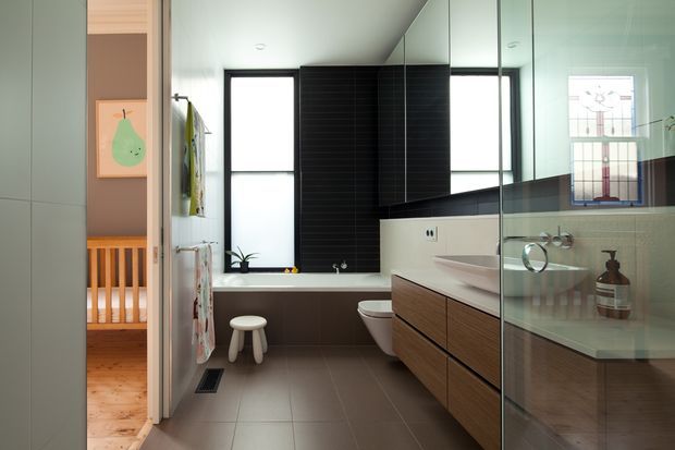 A contemporary family bathroom viewed from the doorway, featuring a long wood-veneer floating vanity with a pale benchtop, integrated basin, wall-hung toilet, bathtub beneath a window, dark horizontal wall tiling, and a frameless glass shower enclosure. A timber-floored bedroom or nursery is visible through the open doorway.