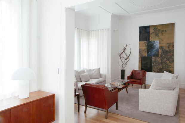 A bright, refined living room viewed through a wide opening, featuring white walls and sheer curtains, a pale upholstered sofa and armchair, two red leather lounge chairs, a glass-topped coffee table, a timber credenza, sculptural decor, and a large abstract artwork. The room has traditional architectural detailing including decorative ceiling molding and wall trim, balanced with mid-century-inspired furniture.