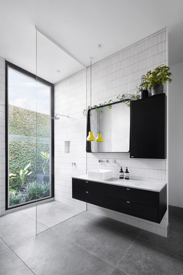 Contemporary bathroom with a large frameless glass shower enclosure, floor-to-ceiling white grid-pattern wall tiles, large grey floor tiles, and a floating matte-black vanity with a white vessel basin. A wide rounded-corner mirror, black wall cabinet, greenery, and bright yellow pendant lights add strong graphic accents. Full-height glazing opens toward a planted courtyard.