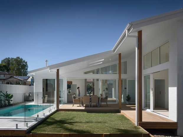 Bright contemporary single-storey residence arranged around a compact central grass courtyard and swimming pool. The white rendered exterior has clean horizontal rooflines, extensive floor-to-ceiling glazing, timber posts and a timber-lined raised deck, with a covered outdoor dining area opening directly to the courtyard.