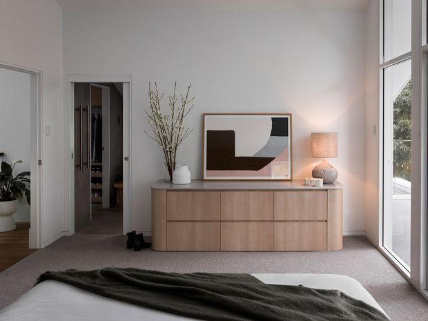 A softly lit contemporary bedroom with a low-profile bed in the foreground, warm timber built-in sideboard cabinetry, neutral carpet, and a large abstract artwork. Full-height glazing along the right side brings in daylight and views of greenery. An open walk-in wardrobe is visible through the doorway, with a small plant-filled adjacent space at left. The room has a calm, refined, minimalist feel with warm natural materials and muted tones.