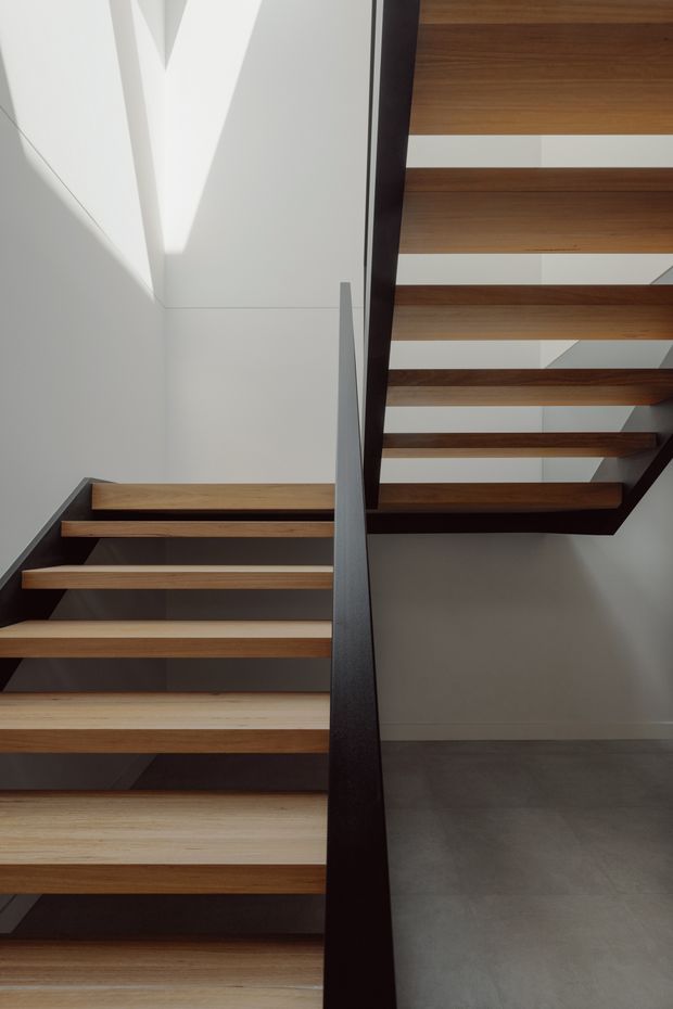 Interior architectural photograph looking up through a modern stairwell. Floating timber stair treads are supported by dark painted steel stringers and a central dark steel partition/guard, with white plaster walls and glimpses of the upper flight creating strong geometric lines and shadows. A smooth grey floor is visible below.