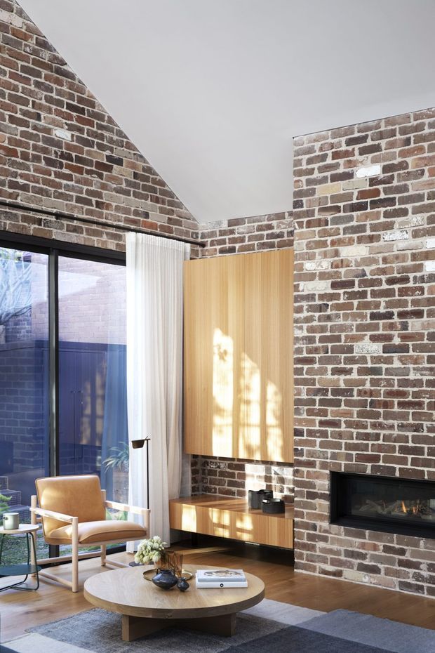 A contemporary living room with exposed red-brown brick walls, a high angled white ceiling, and full-height black-framed glazing with sheer curtains. The room features a warm timber media cabinet and vertical timber panel, a built-in black fireplace, a tan leather lounge chair, a round timber coffee table, and a muted grey rug. Natural light creates a warm, refined atmosphere.