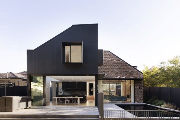A rear exterior view of a contemporary multi-level residence combining a dark, sharply articulated vertical extension with an existing exposed brick volume and hipped roof. Large glazed openings connect the open-plan kitchen and dining area to a timber deck, with a lower terrace and metal balustrade in the foreground. The image highlights the contrast between the black-clad addition, brickwork, extensive glazing, and outdoor living spaces.