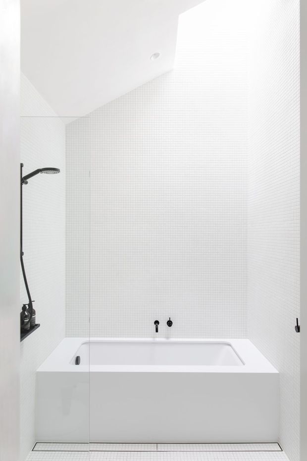 Bright minimalist bathroom featuring a built-in white bathtub set into a tiled surround, a frameless glass shower screen, black shower and tapware, and small white grid-pattern wall and floor tiles. The space has a clean, high-key monochromatic appearance with a plain white ceiling and recessed downlight.