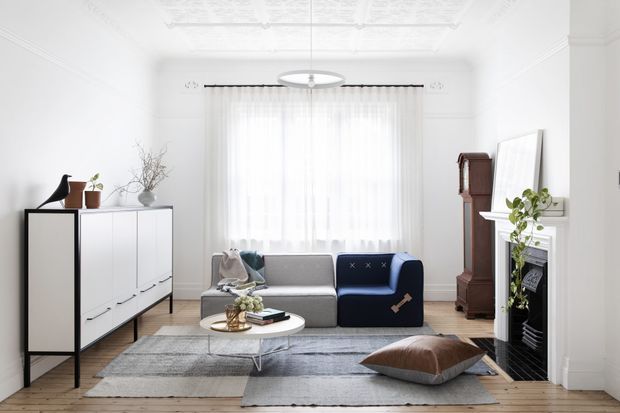 Bright, refined living room with white walls, decorative ceiling detailing, sheer full-height curtains, a grey sofa with a navy throw, layered grey rugs, a round white coffee table, and a traditional fireplace. A white and black sideboard, timber flooring, vintage grandfather clock, plants, and restrained decor create a contemporary interior with classic architectural elements.